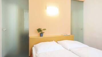 Una camera d'hotel con un letto, un comodino, una pianta in vaso e un punto luce a parete.