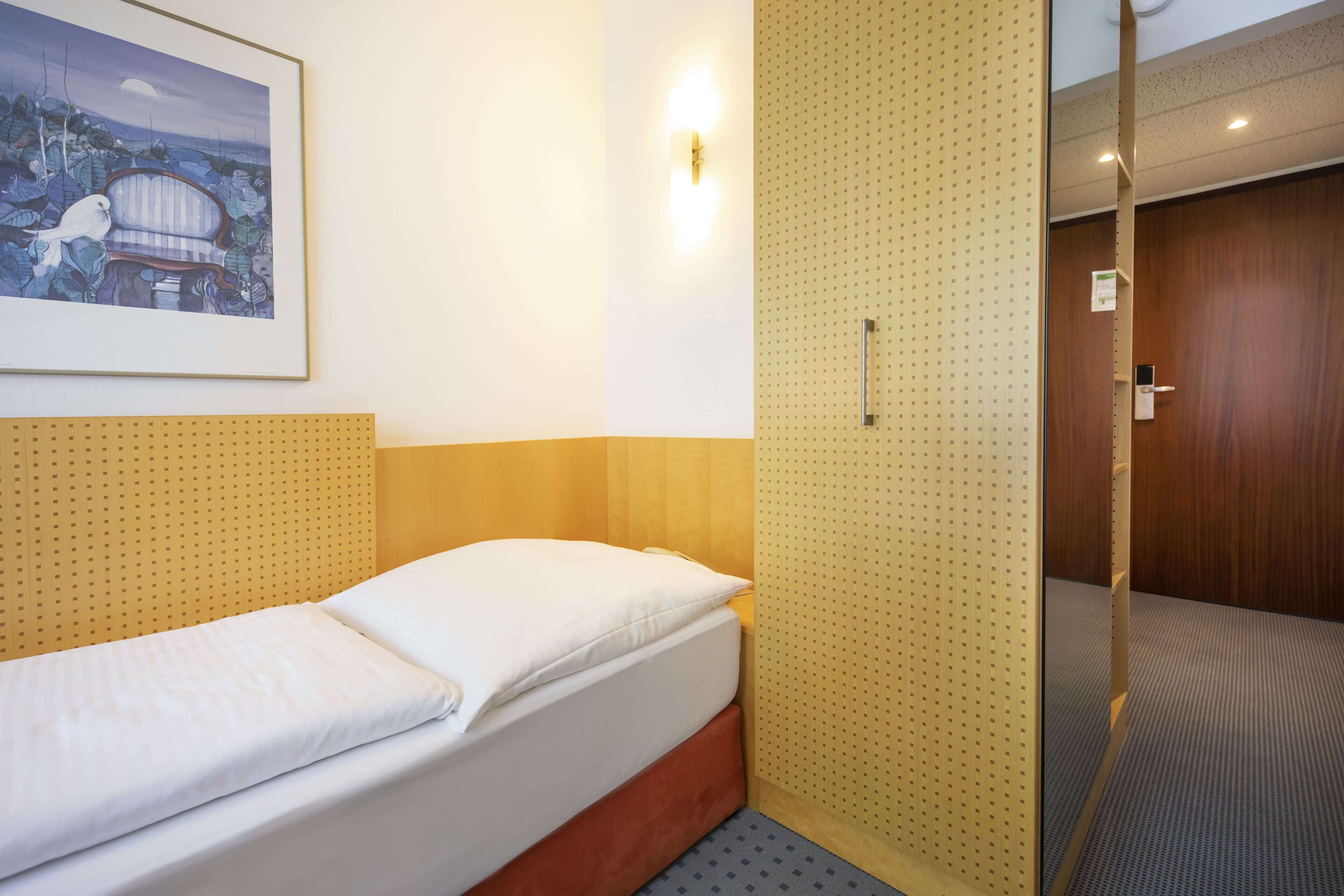 Una cama individual con cabecera de madera y colchón blanco, una lámpara fijada a la pared y un cuadro enmarcado sobre la cama.