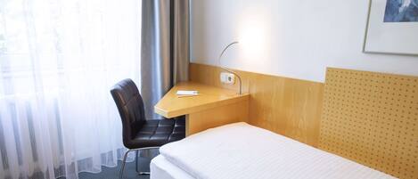 Una habitación de hotel con cama, escritorio, silla y ventana con cortinas traslúcidas.