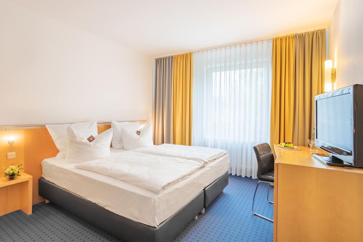 A hotel room with a double bed, a desk with a television, a chair, and a small table with a vase of flowers.