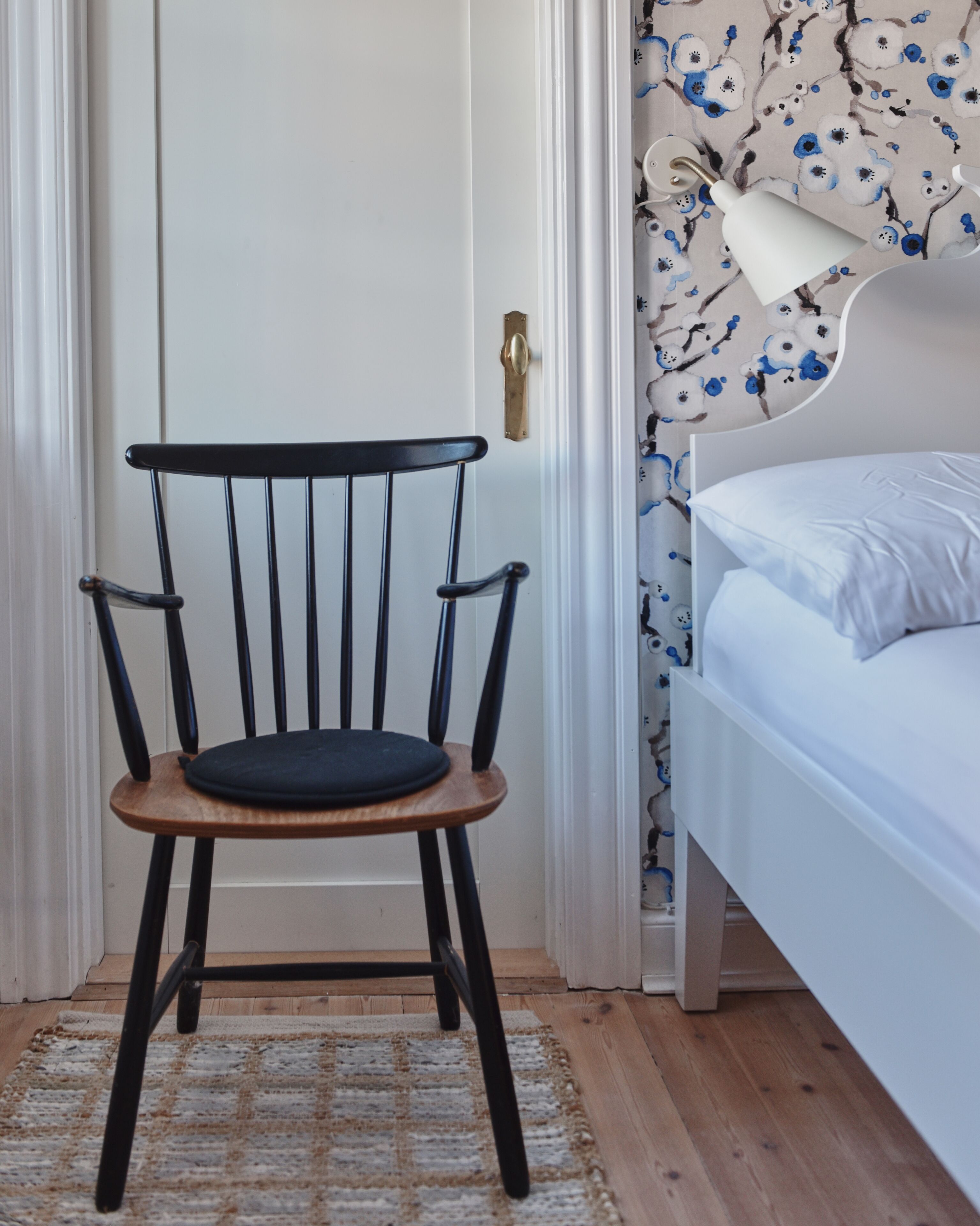 Een kamer met een houten vloer, een wit bed met een wit kussen, een zwarte stoel met een houten zitting, en een wand met een bloemen behangpatroon.