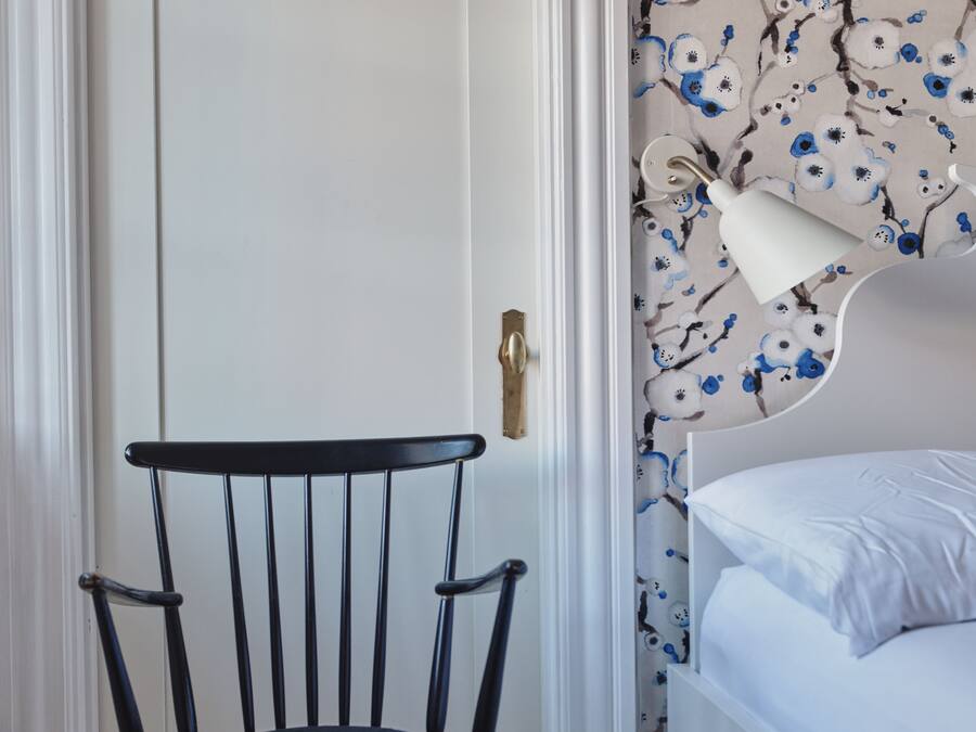 Een kamer met een houten vloer, een wit bed met een wit kussen, een zwarte stoel met een houten zitting, en een wand met een bloemen behangpatroon.