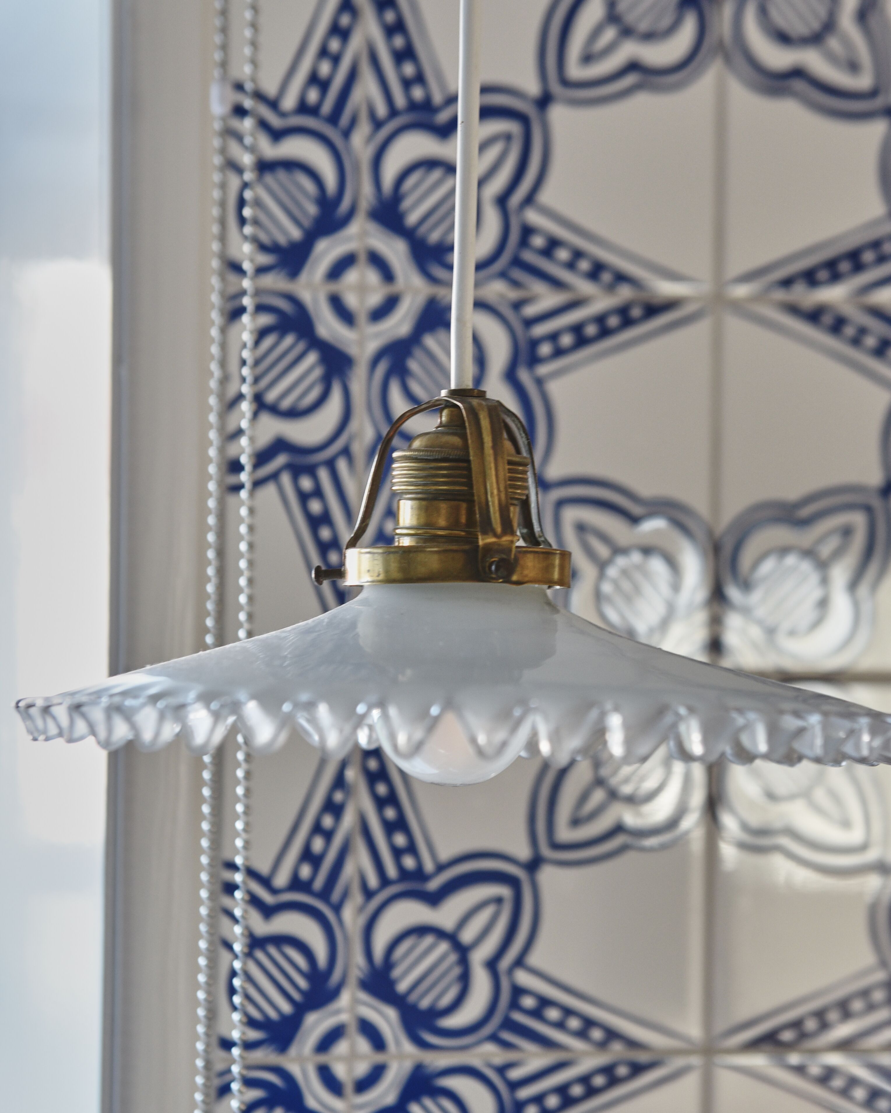 A hanging light fixture with a clear glass shade and a brass base, set against a wall with blue and white patterned tiles.