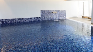A clear indoor swimming pool with a tiled edge, a water feature, and a seating area with loungers. - Strandhotellet - Blokhus (Blokhus)