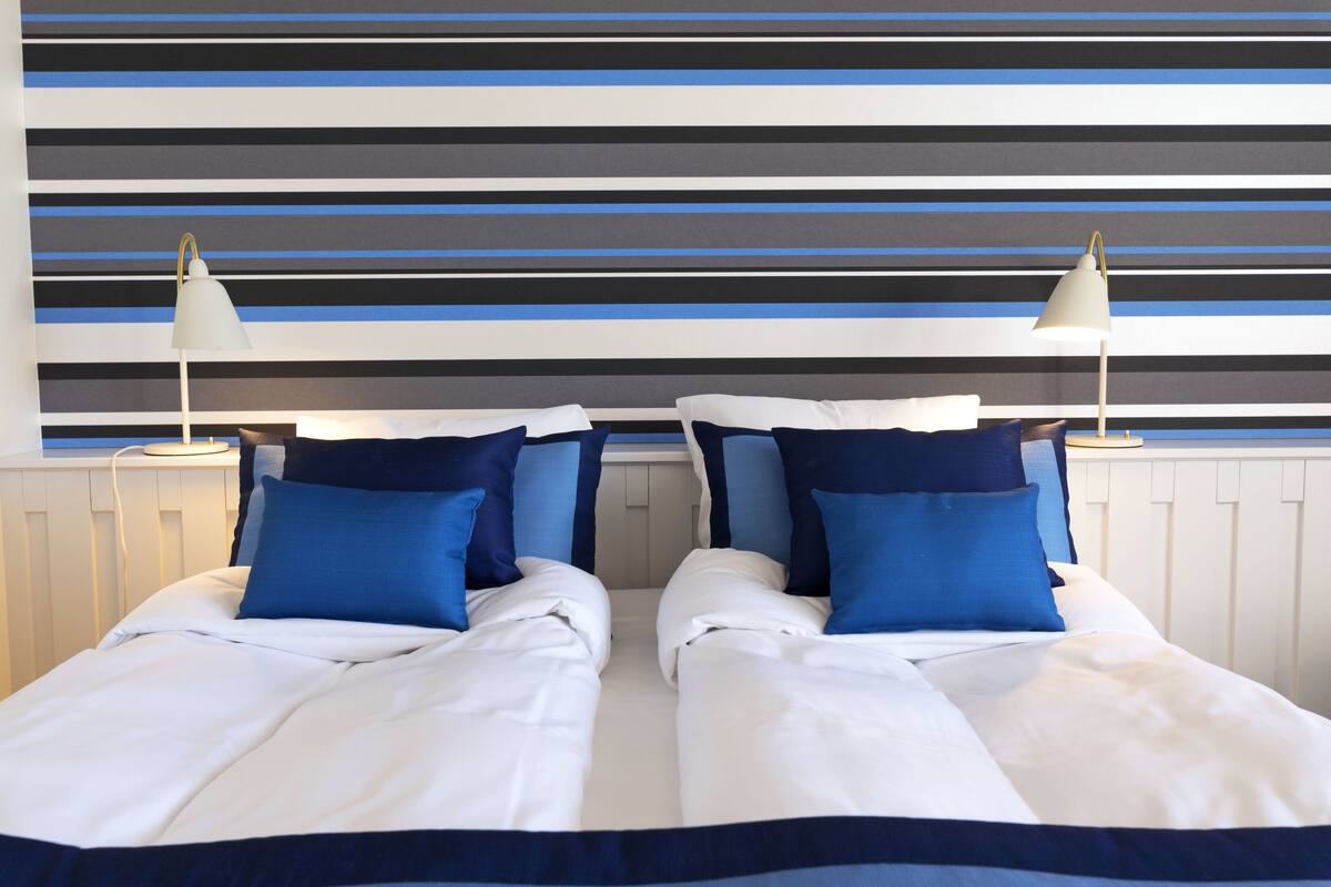 A double bed with white and blue bedding, two bedside lamps, and a striped wall behind.