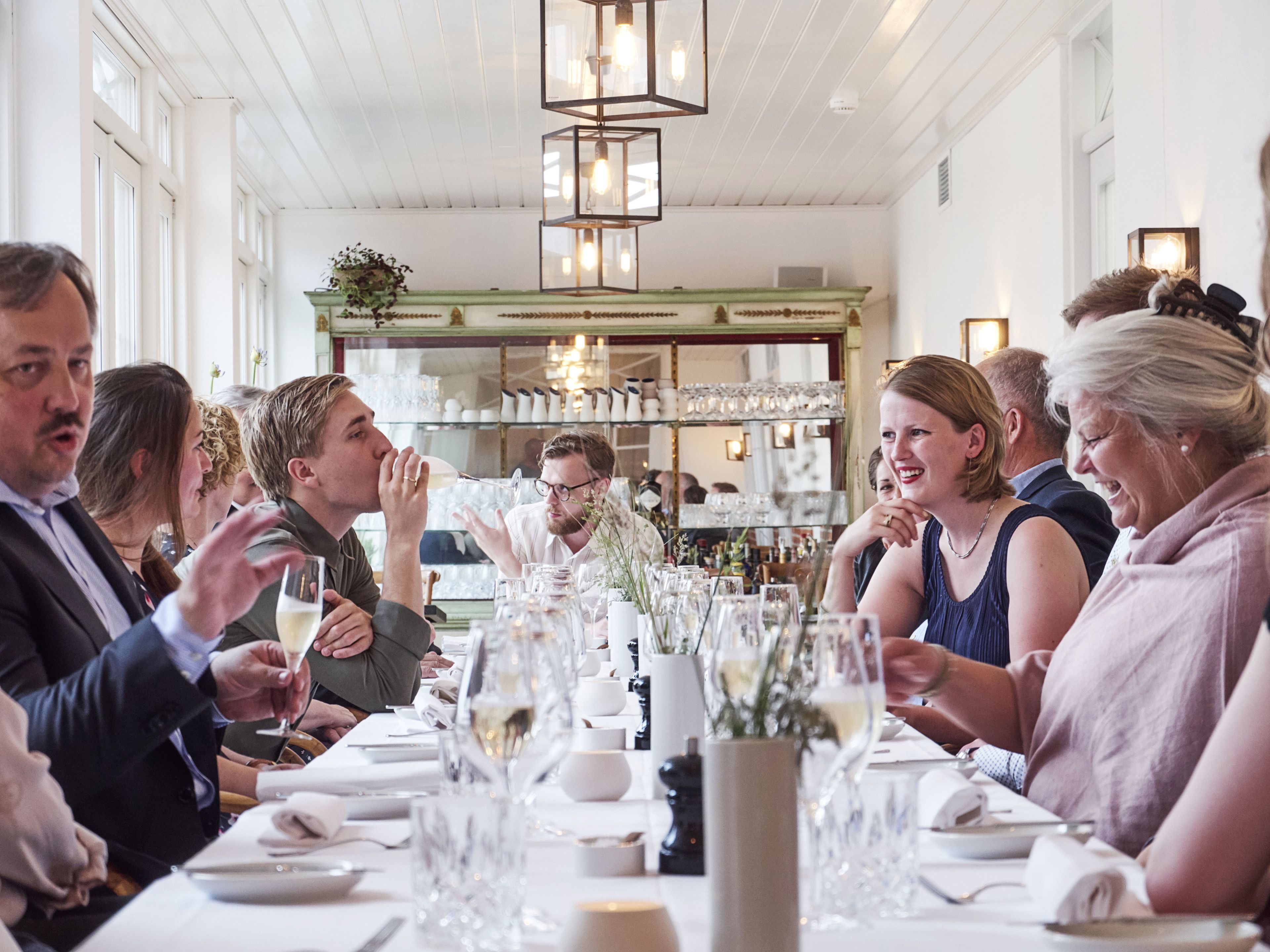 Un grupo de personas cenando en una mesa con manteles blancos, rodeado de copas y botellas, en una sala con paneles de madera y un candelabro.