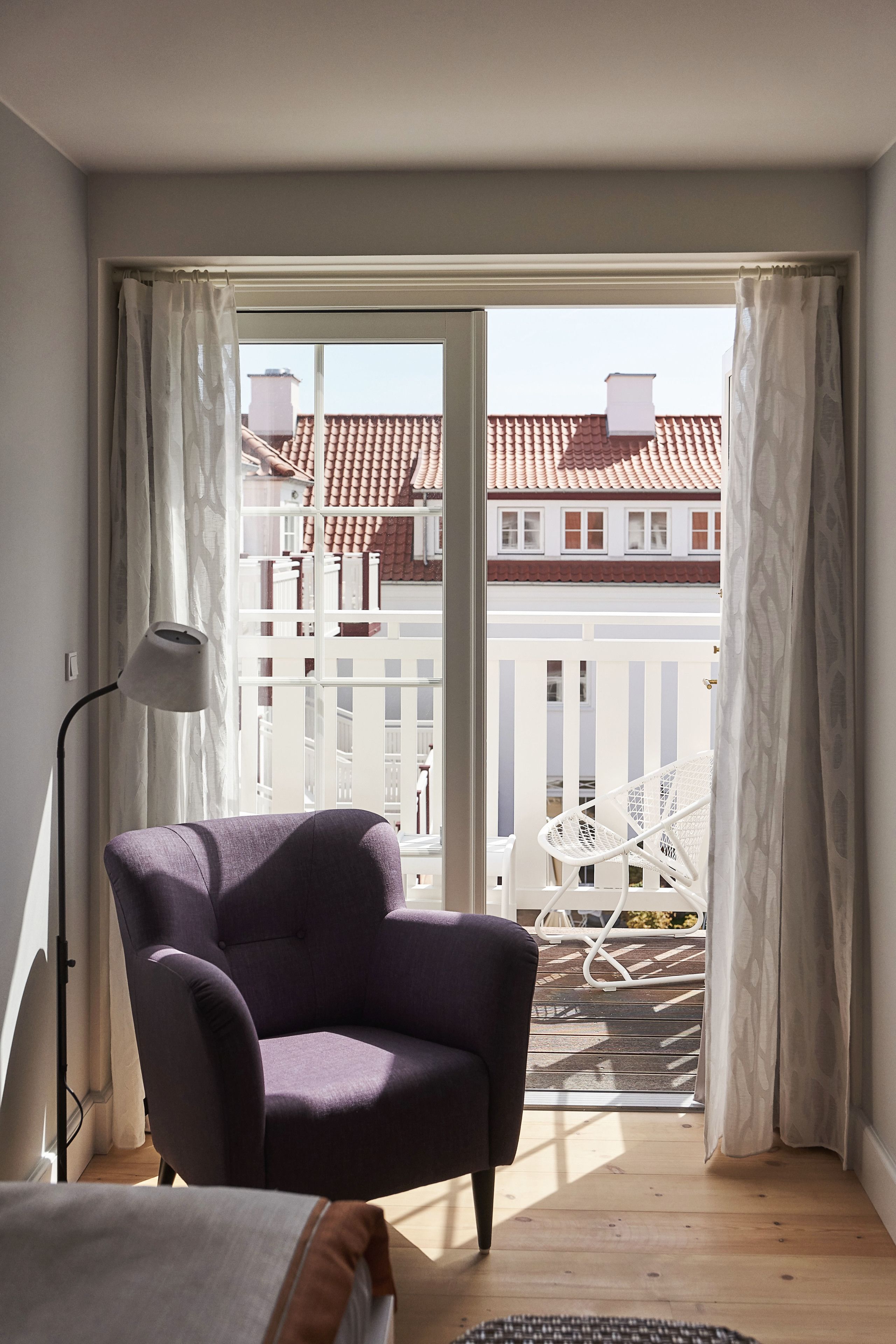 A room with a purple armchair, a floor lamp, and a view of a balcony with a white railing and a red-tiled roof.