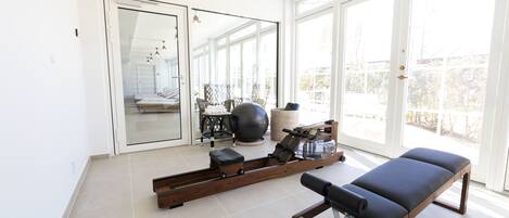 A modern room with a rowing machine, a massage table, and a large mirror reflecting a balcony with outdoor seating.