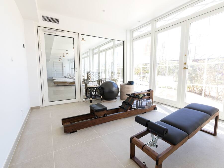 Una habitación moderna con una máquina de remo, una mesa de masajes y un espejo grande que refleja un balcón con mobiliario exterior.