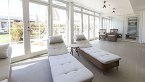 A bright, sunlit room with large sliding glass doors leading to a patio, featuring a long white sofa and wicker chairs. - Strandhotellet - Blokhus (Blokhus)