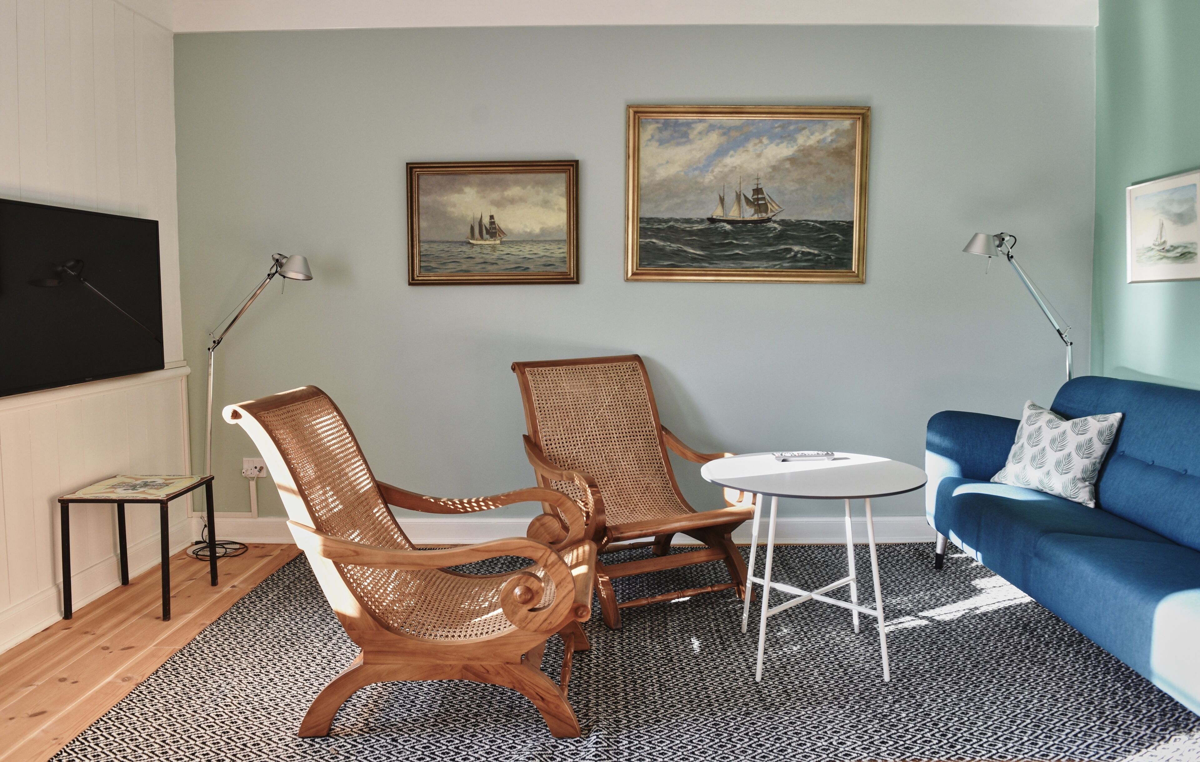 A living room with two wooden chairs, a small round table, a blue sofa, and two paintings on the wall.