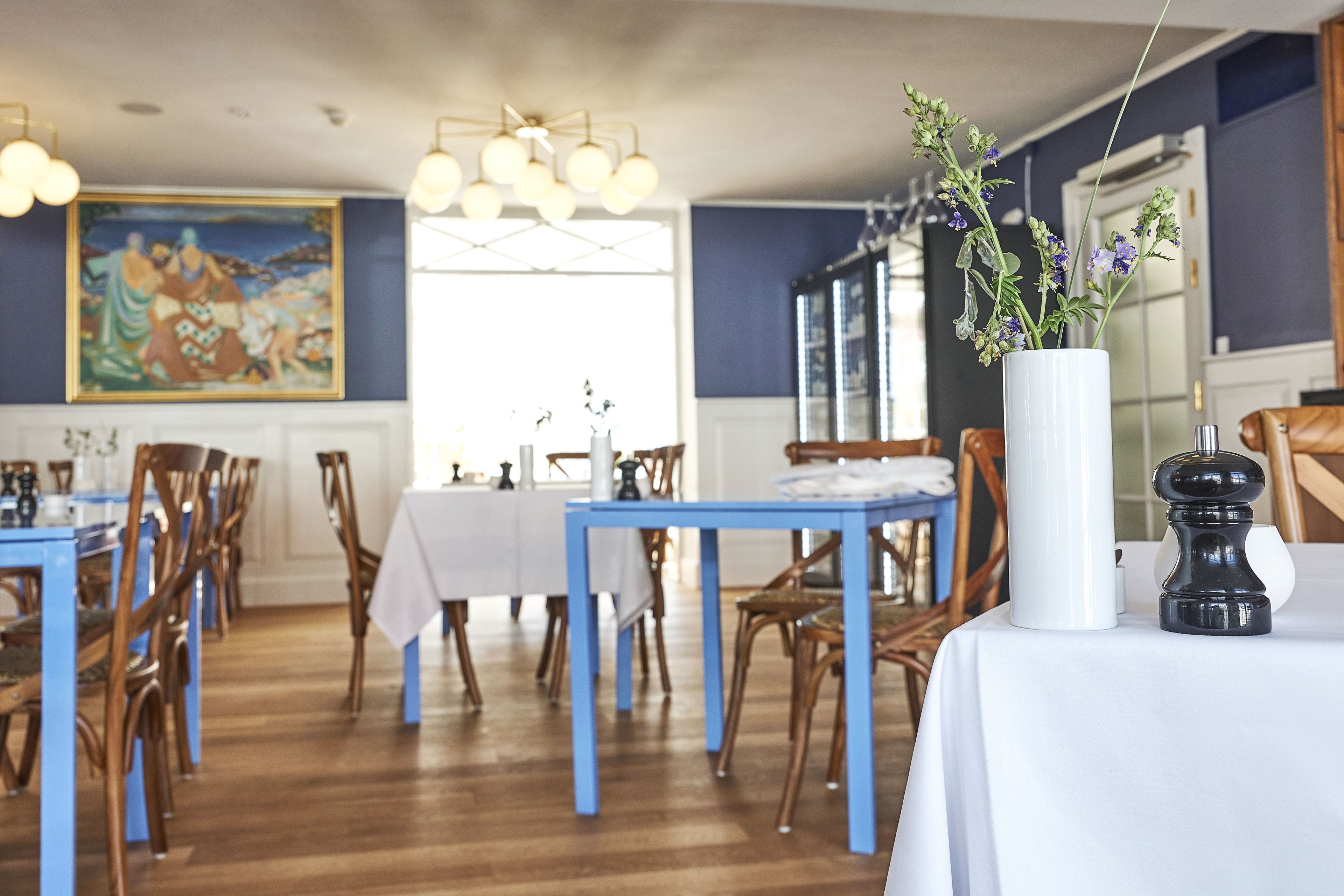 A dining area with blue tables and chairs, a white vase with flowers, and a painting on the wall.