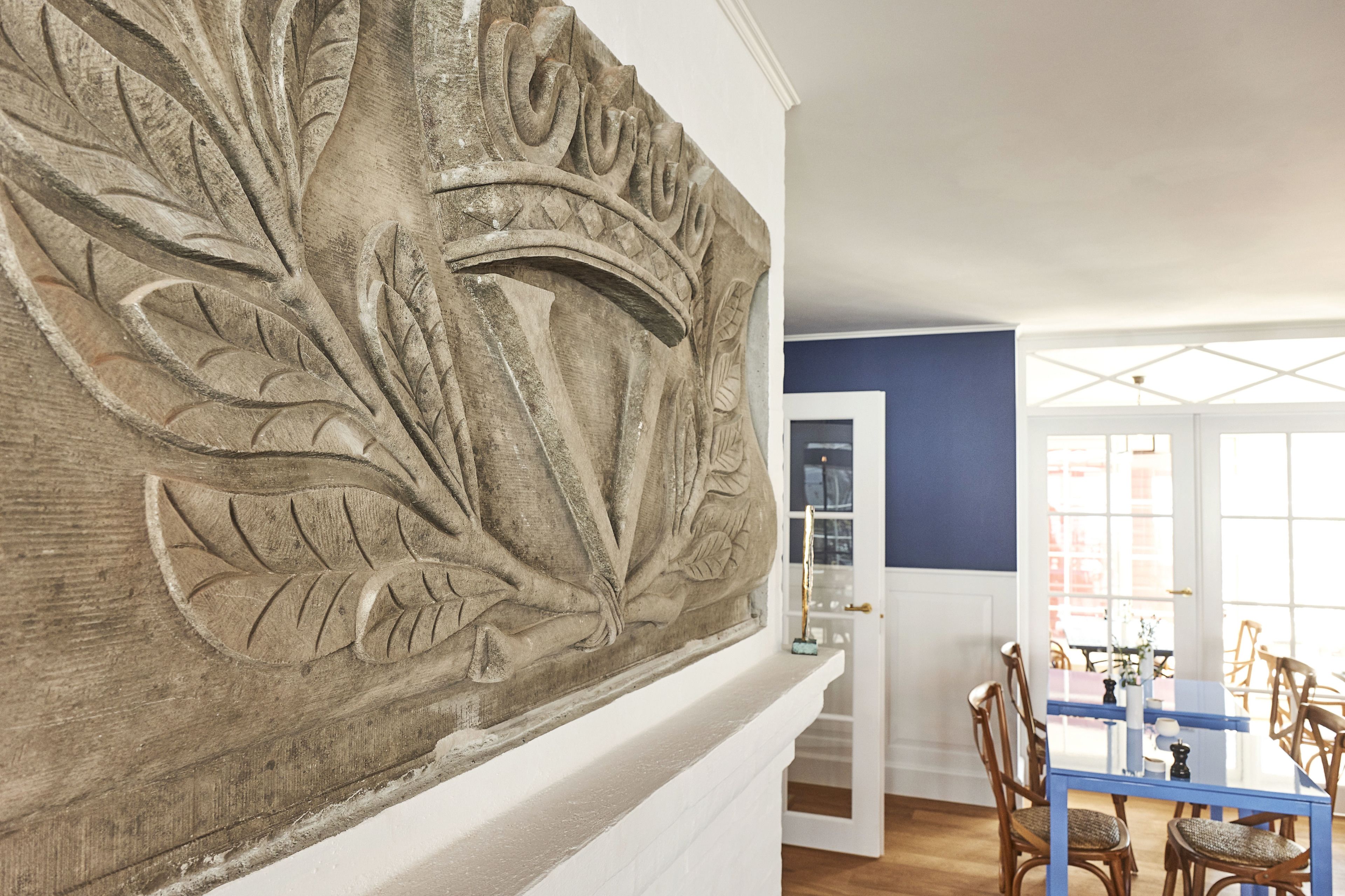 A dining area with a blue table and wicker chairs, a white fireplace with a carved stone mantle, and a view through a glass door to the outside.
