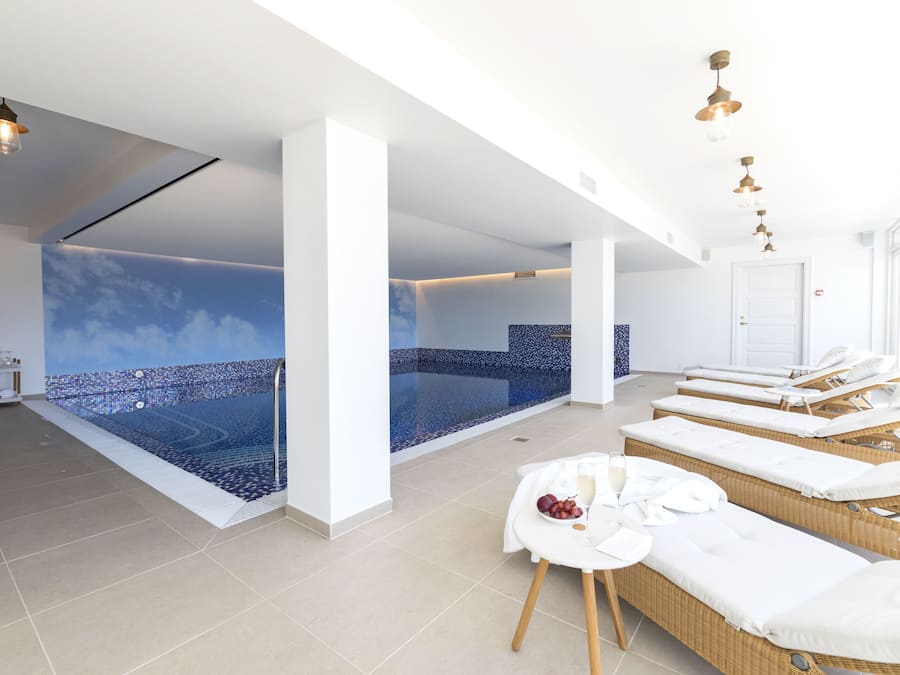 Un spa moderno con una amplia piscina interior, tumbonas y una pequeña mesa con frutas y toallas.