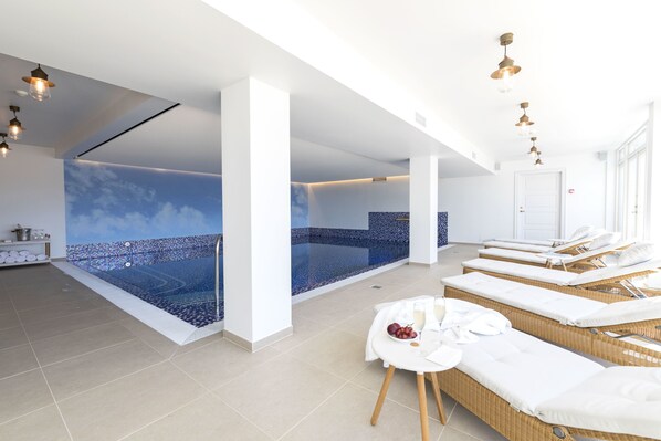 A modern spa room with a large indoor pool, sun loungers, and a small table with fruits and towels.