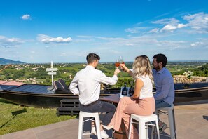 Three people seated at an outdoor table with a scenic view, a boat, and a signpost in the background. - Hotel Villa del Poggio (San Pietro di Feletto)