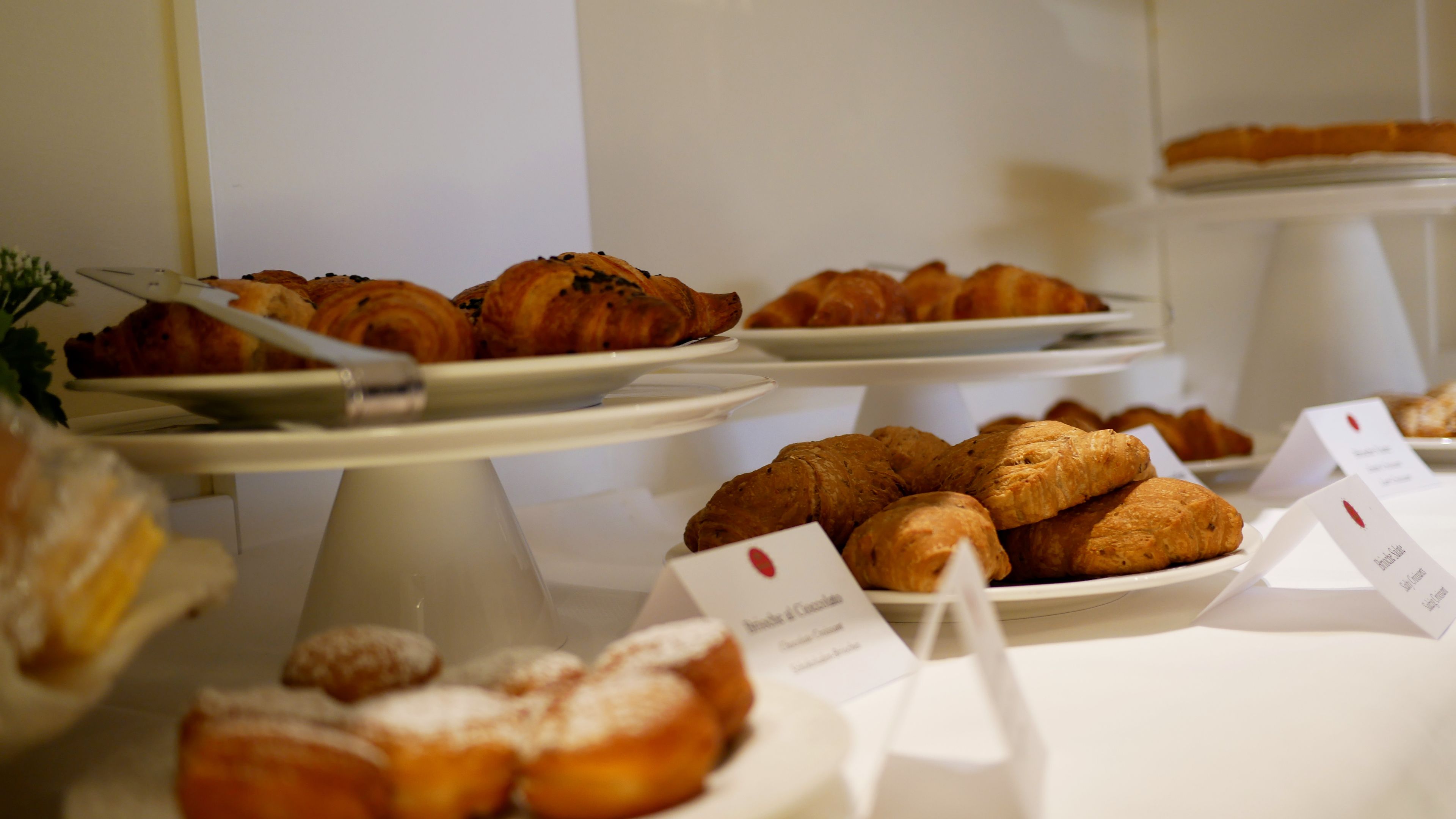 A buffet with various pastries and desserts, including croissants and donuts, displayed on white stands with labels.