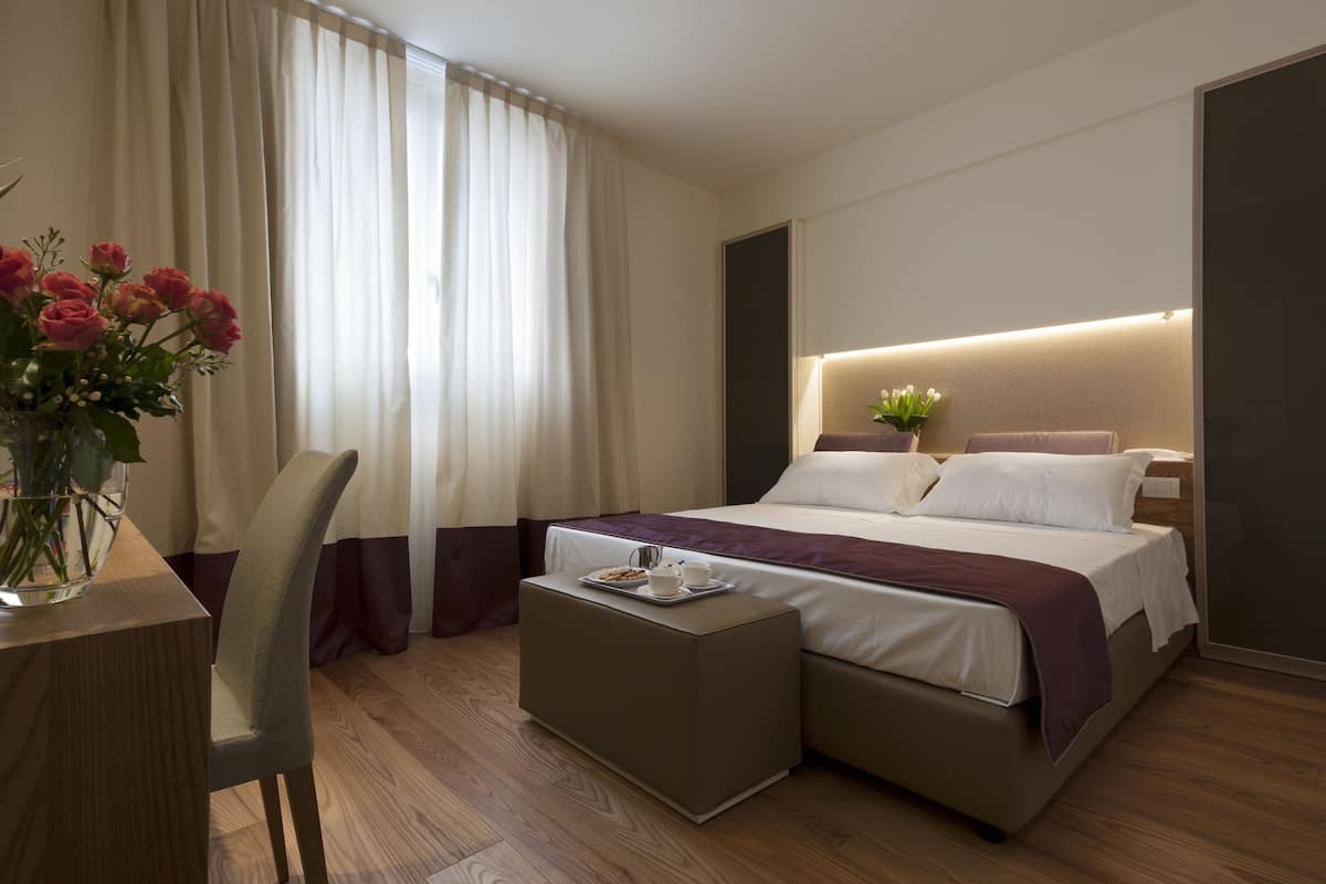 Ein modernes Hotelzimmer mit einem großen Bett, einem Nachttisch mit einer Kuchensämlung, einer Blumenvase und einem Sessel.
