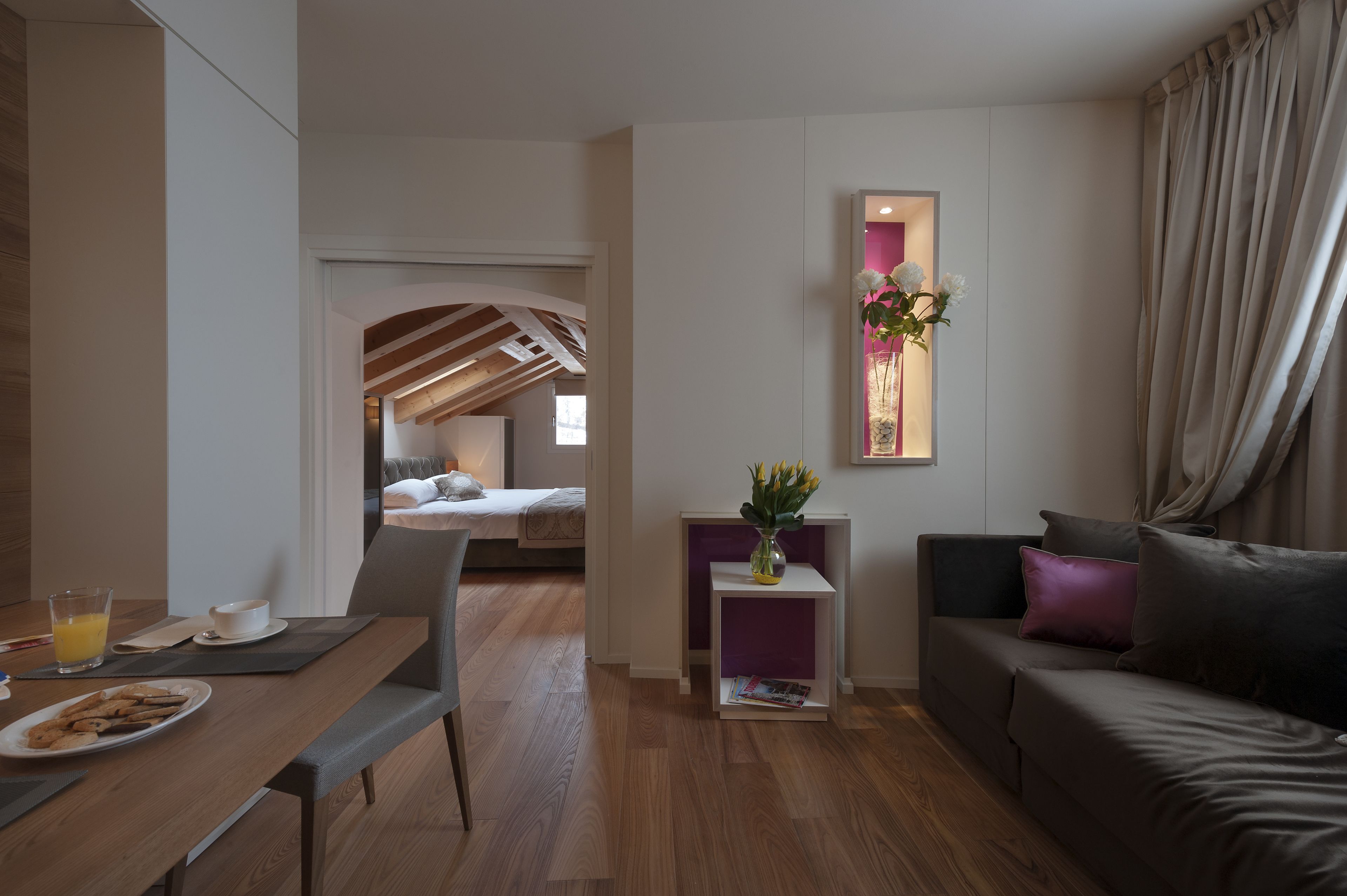 A modern hotel room with a dining area, a sofa, a small table with a vase of flowers, and a view of the bedroom.