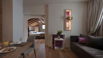 A modern hotel room with a dining area, a sofa, a small table with a vase of flowers, and a view of the bedroom.