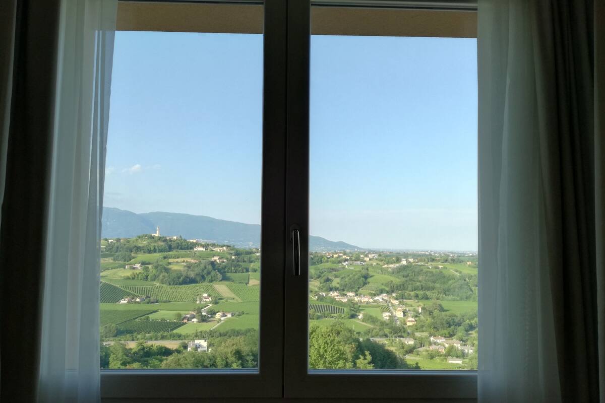 Ein Fenster mit Blick auf eine grüne Landschaft, darunter Felder und ein entferntes Gebäude.