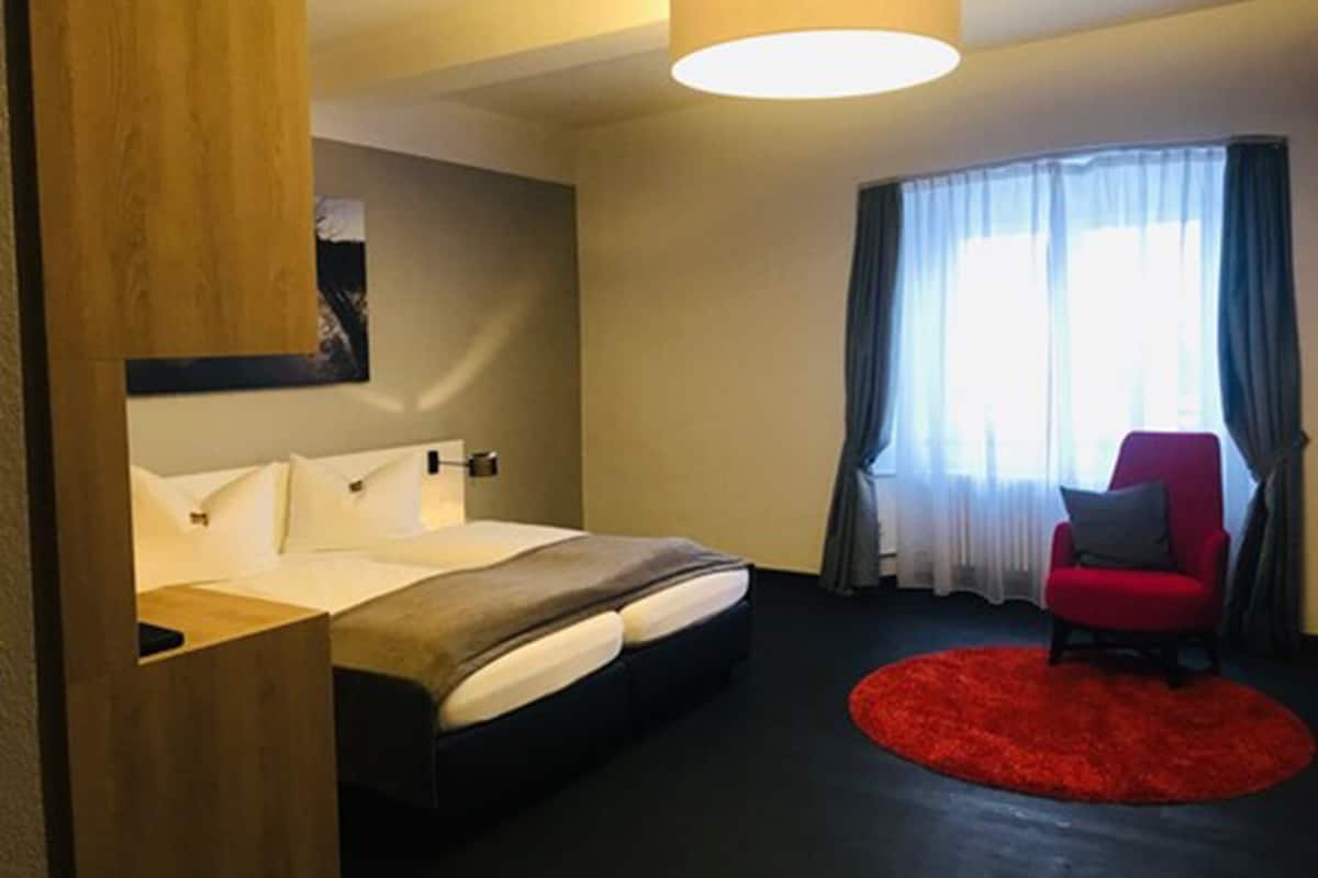 A hotel room with a bed, a red chair, a red rug, and a window with curtains.