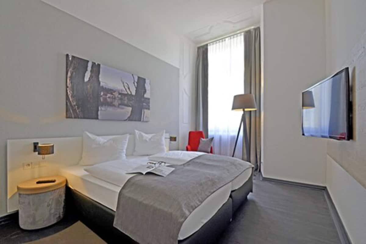 A hotel room with a bed, a bedside table, a lamp, a TV, and a window with curtains.