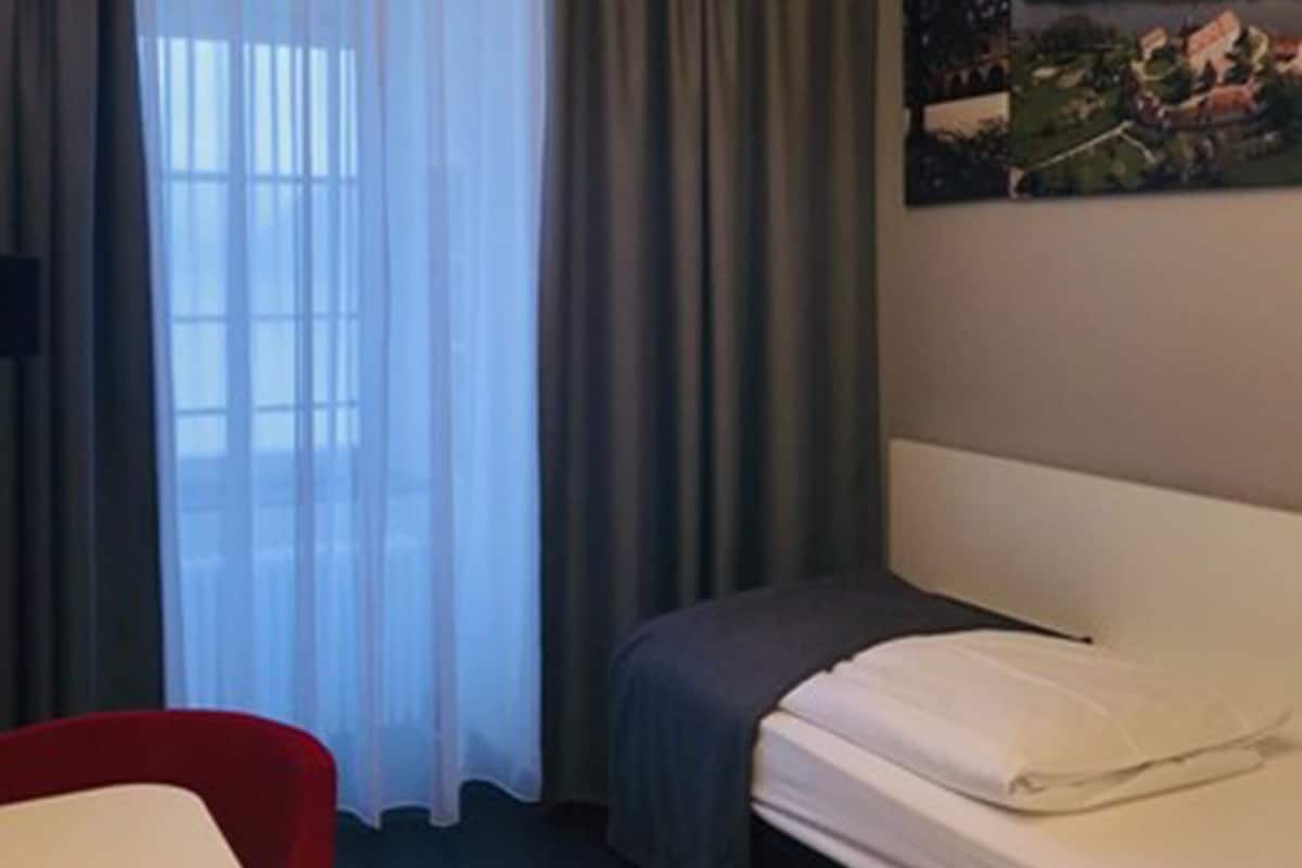 A hotel room with a bed, a desk, a chair, a painting on the wall, and a window with curtains.