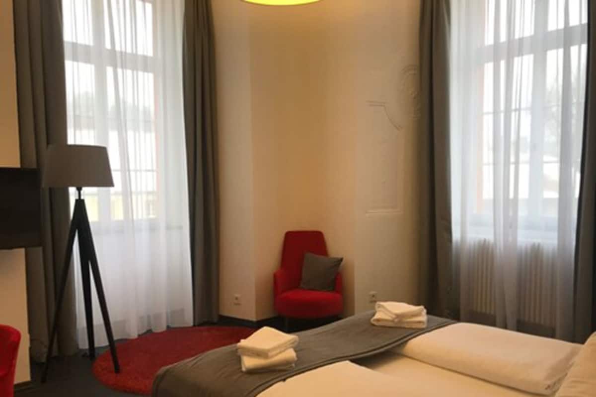 A hotel room with a bed, a red chair, a floor lamp, and a view through large windows.