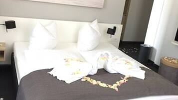 A neatly made bed with white linens and a floral arrangement, a painting on the wall, and a trash bin to the side.