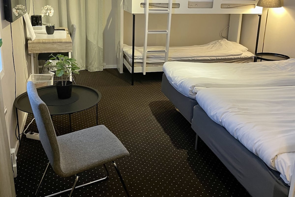 A hotel room with bunk beds, a desk with a chair, a small table with a plant, and a lamp.