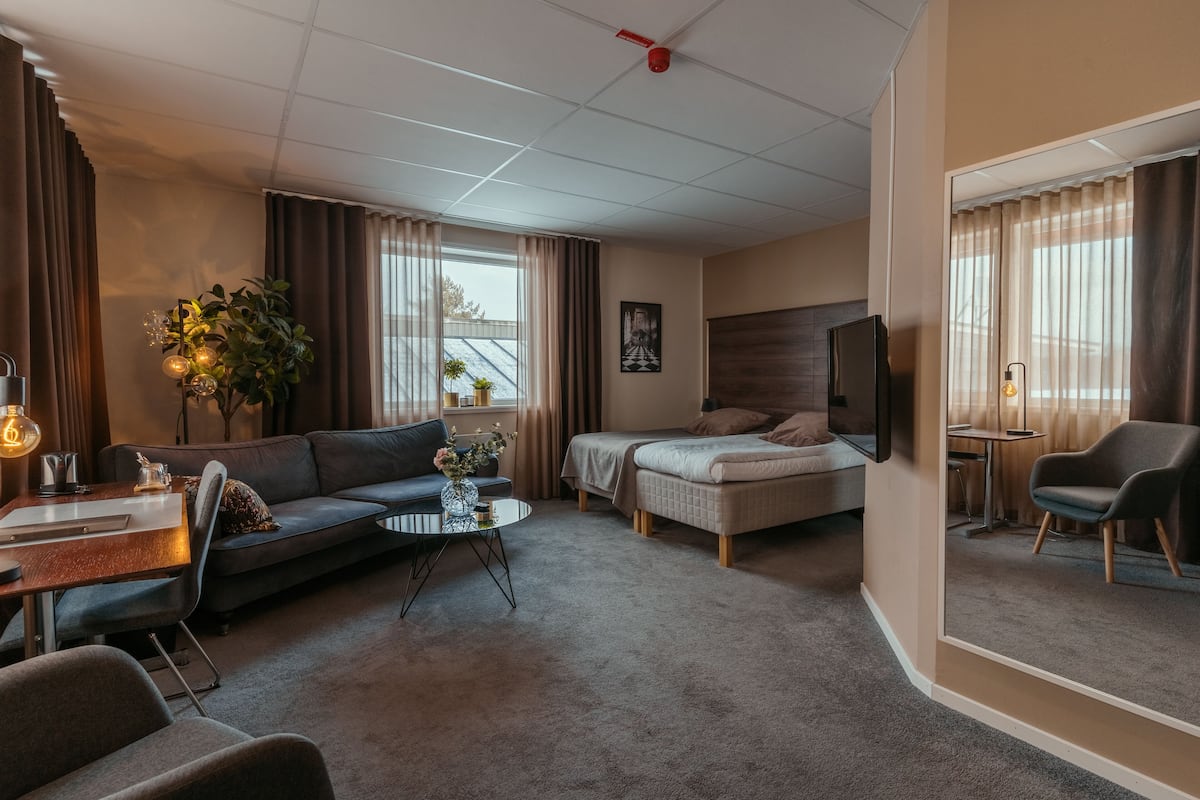 A modern hotel room with a sofa, a bed, a desk, and a chair. There is a plant, a lamp, and a mirror.