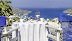 A table set with a white tablecloth, two wine glasses, a pitcher, and a small vase with purple flowers, overlooking a coastal view. - Skopelitis Village (Amorgos)