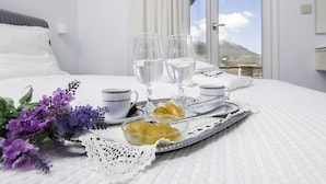 A neatly arranged breakfast tray with fruits, glasses, and cups on a white bedspread, with a mountain view through the window. - Skopelitis Village (Amorgos)