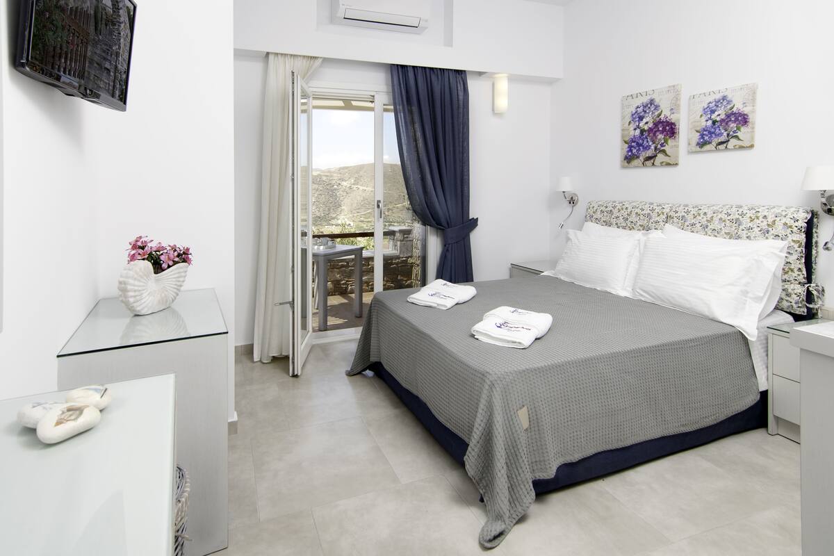 A bedroom with a bed, a nightstand, a dresser, a TV, a balcony with a view, and two paintings on the wall.