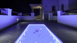 A rooftop terrace with a rectangular illuminated floor feature, a pergola, and a pathway leading to a building entrance. - Skopelitis Village (Amorgos)