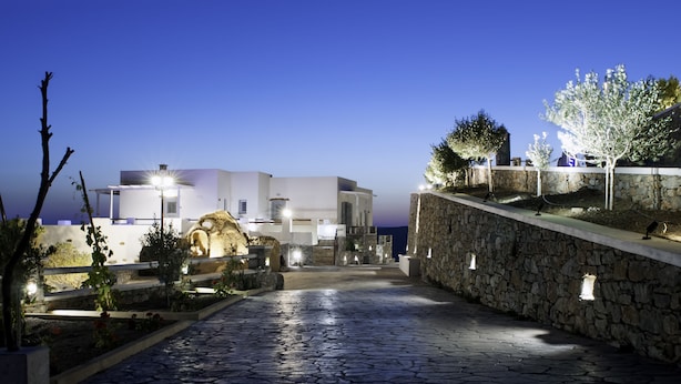 A stone pathway leading to a white building with multiple illuminated windows and a stone wall on the right. - Skopelitis Village (Amorgos)