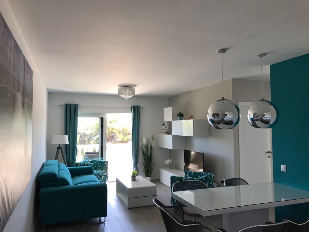 A modern living room with a sofa, a dining table, and a TV. There is a window with curtains and a chandelier.