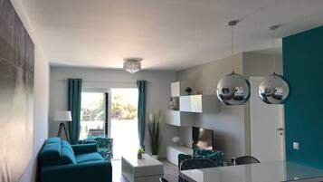 A modern living room with a sofa, a dining table, and a TV. There is a window with curtains and a chandelier.