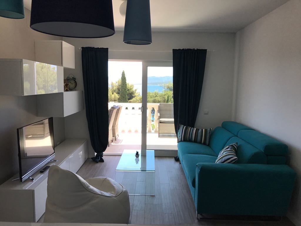 Een moderne woonkamer met een blauwgroene bank, een flatscreen-televisie en een glazen salontafel. Er is een schuifpui die leidt naar een buitenruimte met uitzicht op bomen en bergen.
