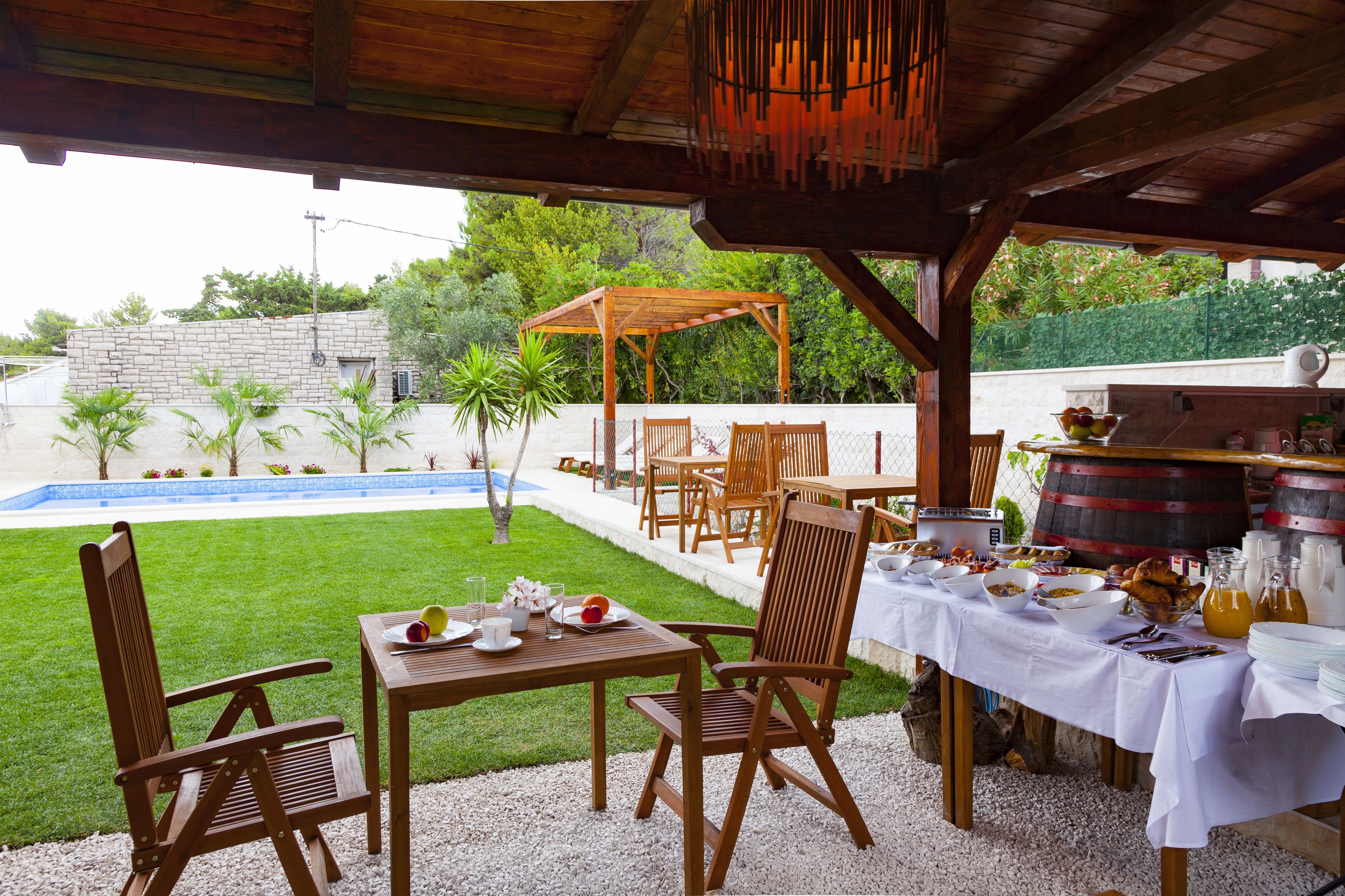 An outdoor dining area with wooden tables and chairs, a barbecue grill, and a pool in the background.