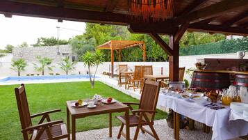 An outdoor dining area with wooden tables and chairs, a barbecue grill, and a pool in the background.