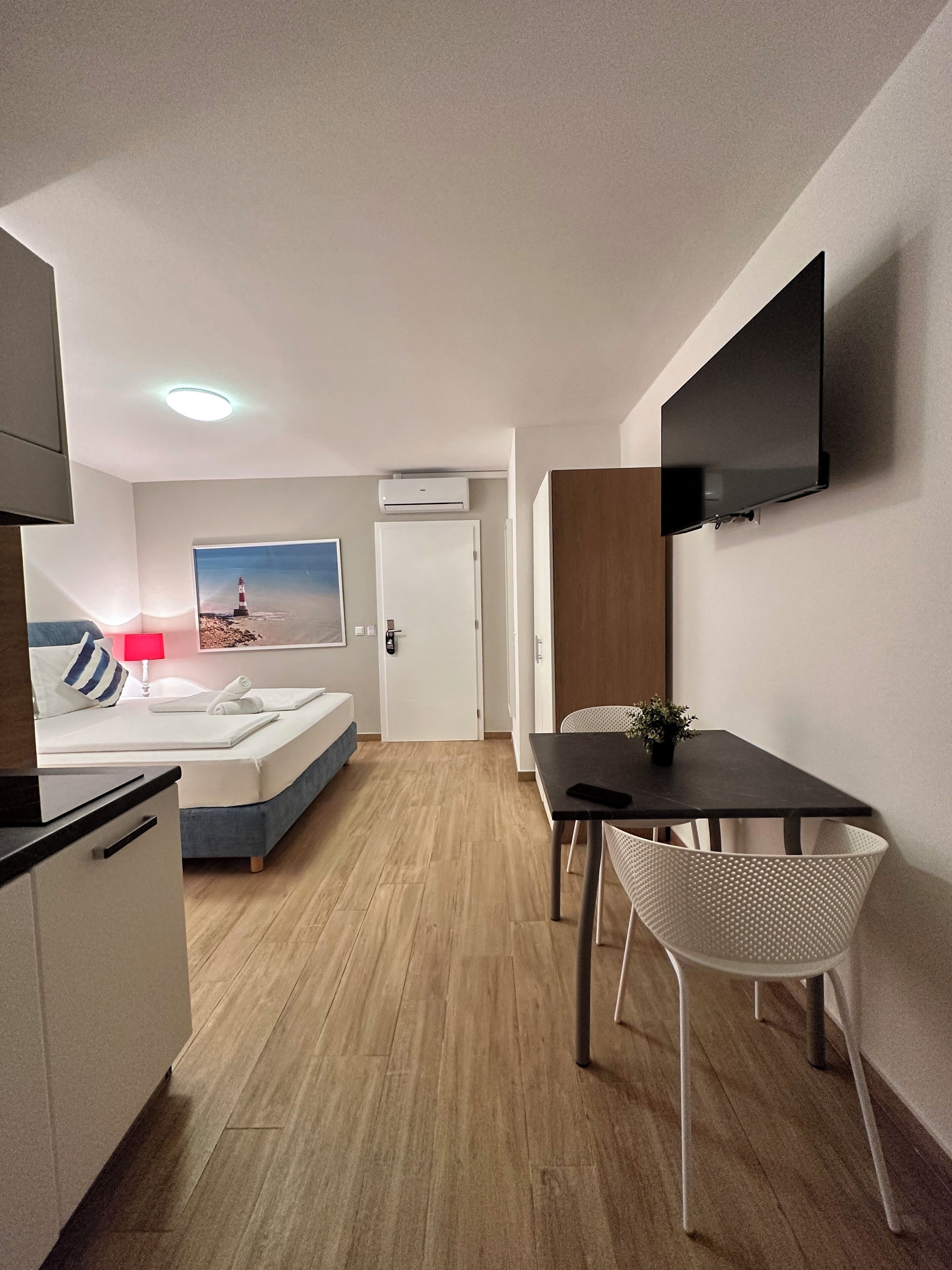 A compact hotel room with a bed, a small kitchenette, a dining area with a table and chairs, a mounted TV, and a painting of a lighthouse on the wall.