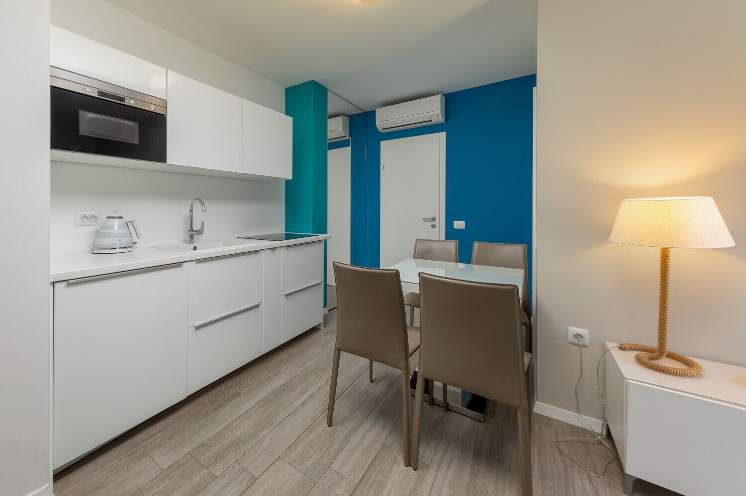 A compact kitchen with white cabinets, a built-in oven, and a coffee maker. There is a dining area with a table and chairs, and a lamp on a side table.