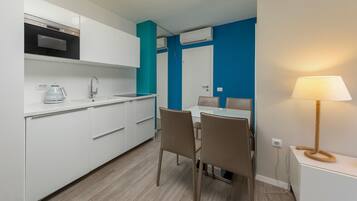 A compact kitchen with white cabinets, a built-in oven, and a coffee maker. There is a dining area with a table and chairs, and a lamp on a side table.