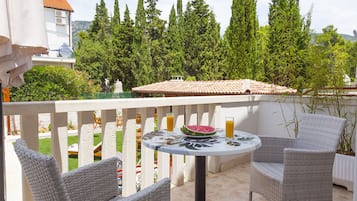 Un balcon avec des fauteuils en osier, une table avec une pastèque et des boissons, et une vue sur des arbres et un bâtiment avec un toit en tuiles.