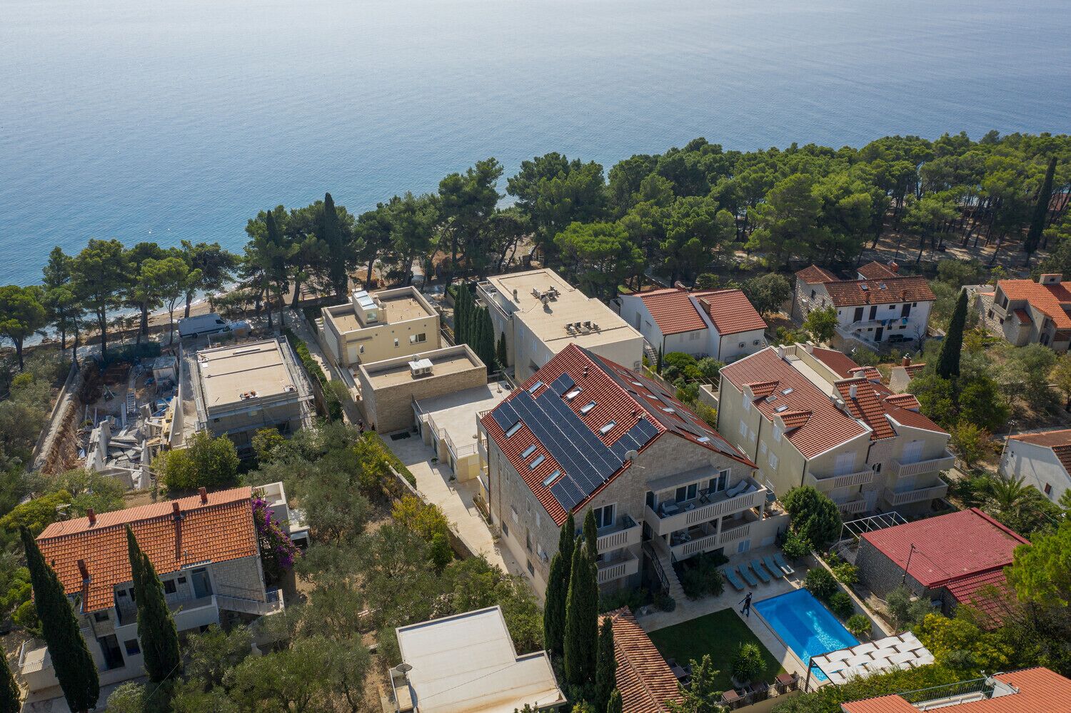 An aerial view of a coastal residential area with houses featuring red-tiled roofs, a swimming pool, and a backdrop of the sea and dense greenery.
