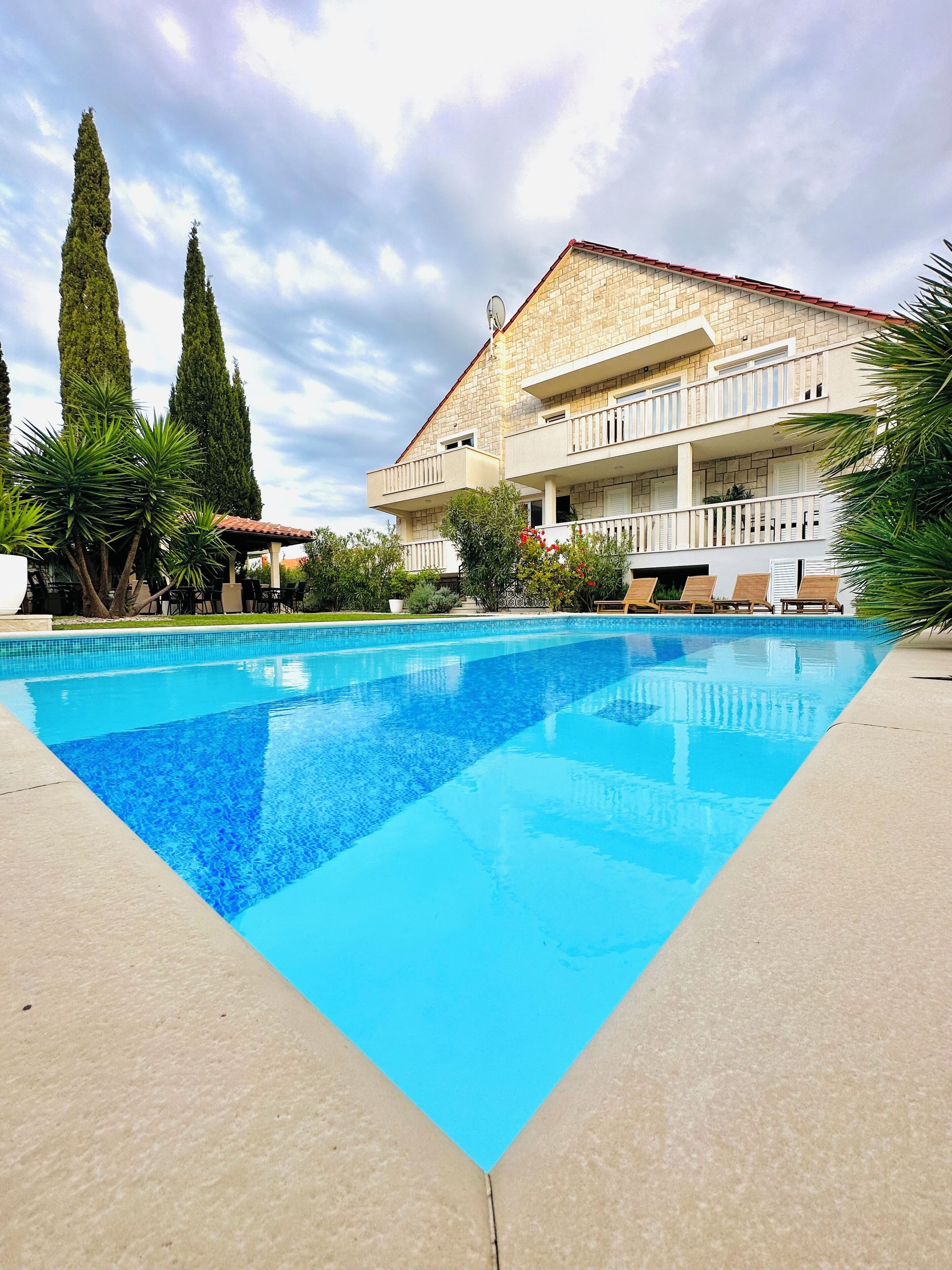 Una piscina con un borde de baldosas, un edificio de dos pisos con balcones y altos cipreses al fondo.