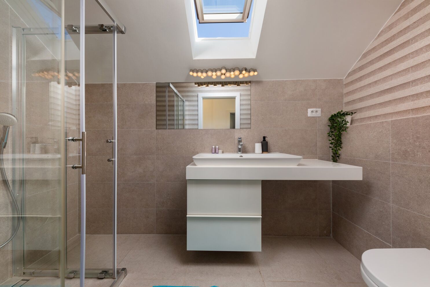 Une salle de bain moderne dotée d’un puits de lumière, d’une cabine de douche en verre, d’un grand lavabo avec un miroir et de toilettes.