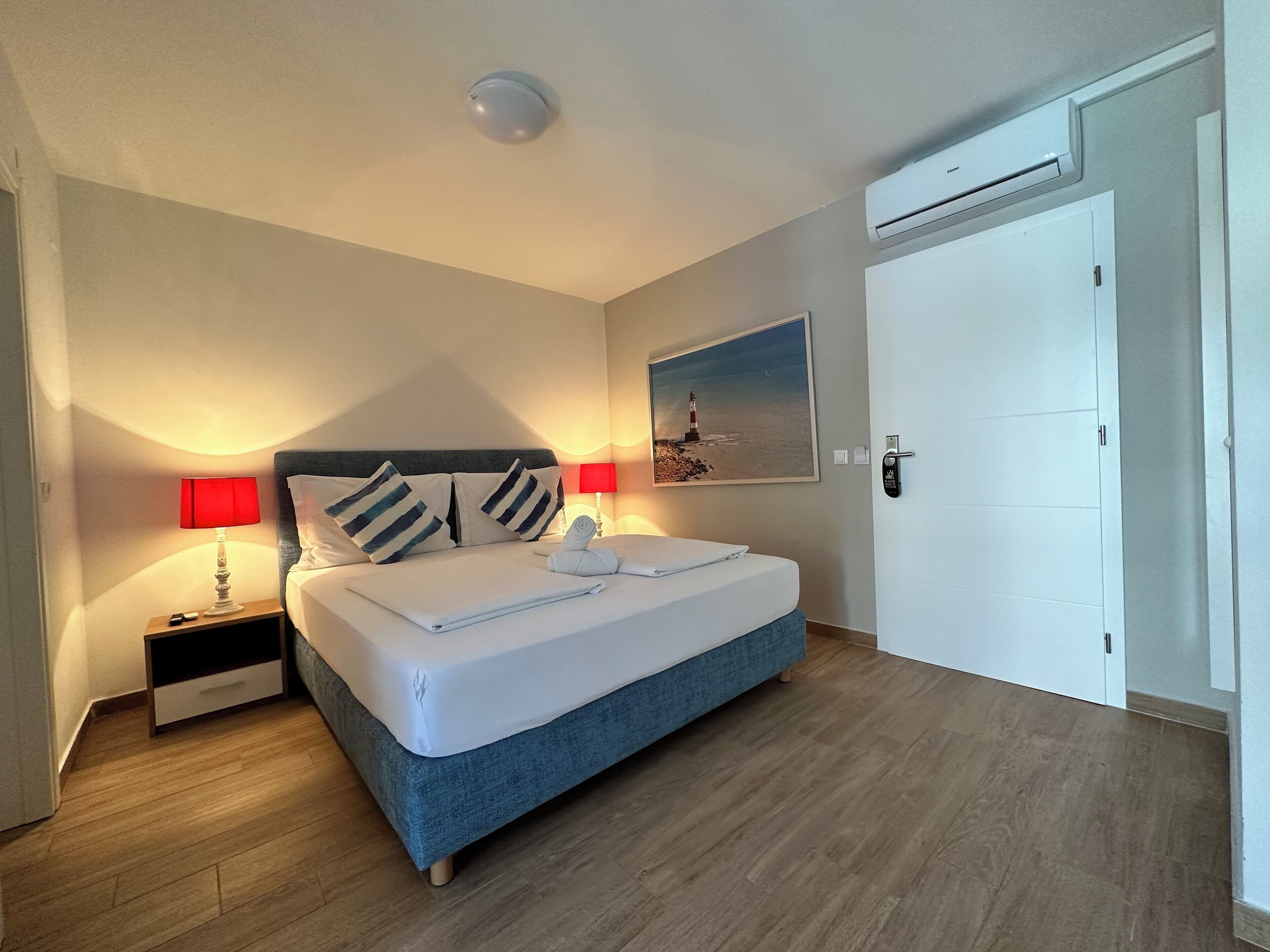 A neatly made bed with a blue headboard, two bedside lamps, a wall-mounted air conditioner, and a door with a handle.